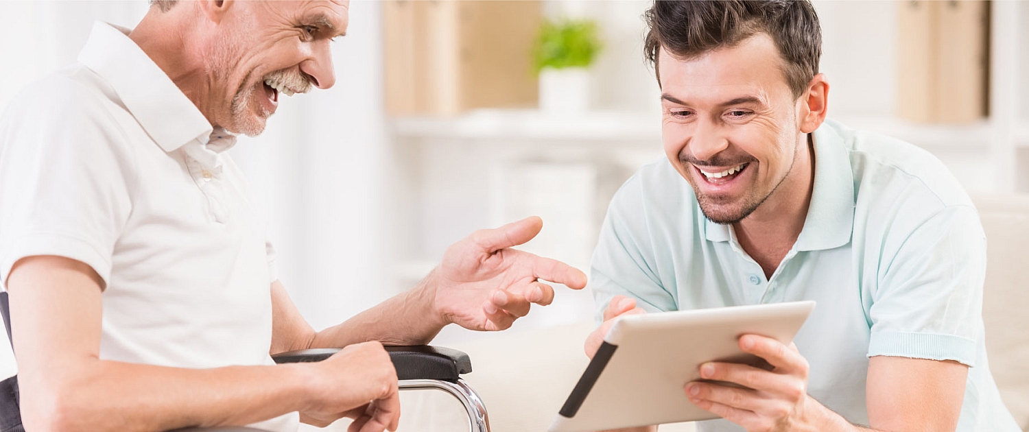 Aide au handicap : un jeun homme montre quelque chose sur un tablet à un ancien assis sur un fauteuil roulant